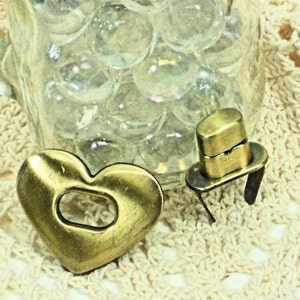 May include: Two brass-toned metal charms: a heart with a rectangular cutout and a miniature top hat. The heart charm is on the left, and the top hat charm is on the right. The background includes a glass jar filled with clear marbles and a cream-colored crocheted surface.