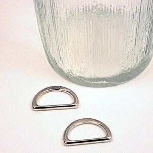 Puede incluir: Dos anillos en D plateados, tambi&eacute;n conocidos como anillos en D, est&aacute;n sobre una superficie blanca. Los anillos tienen un di&aacute;metro de aproximadamente 2,5 cm.