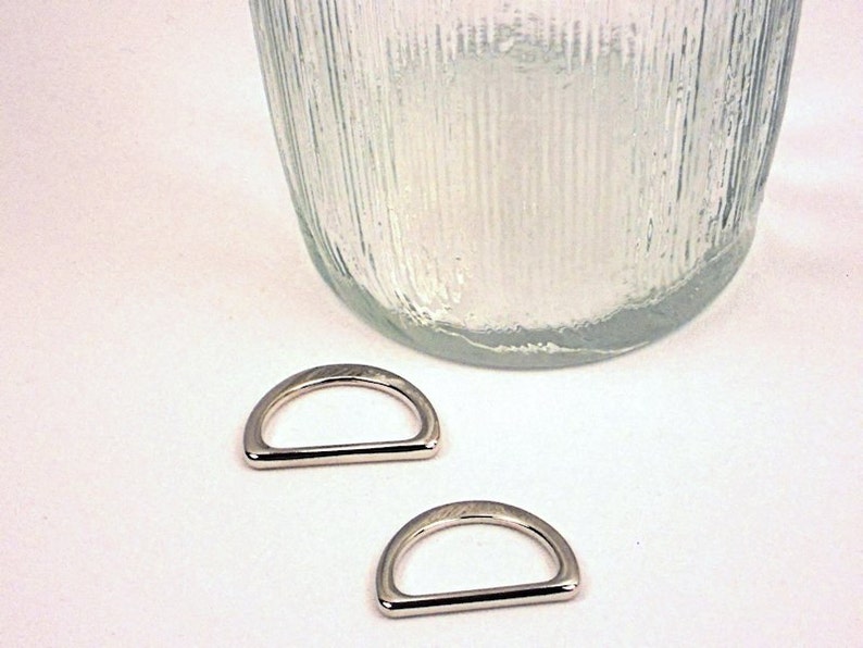 Puede incluir: Dos anillos en D plateados, tambi&eacute;n conocidos como anillos en D, est&aacute;n sobre una superficie blanca. Los anillos tienen un di&aacute;metro de aproximadamente 2,5 cm.