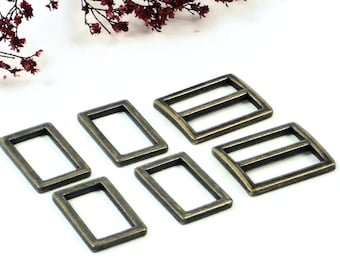 Puede incluir: Seis hebillas deslizantes de metal de color dorado antiguo. Las hebillas son rectangulares con dos agujeros a cada lado. Las hebillas están dispuestas en forma escalonada sobre una superficie blanca.