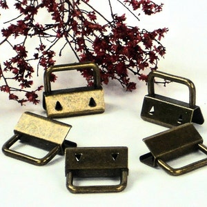 May include: Five antique gold-toned metal slider buckles with a rectangular shape and a small handle. The buckles have a textured finish and are arranged in a circle on a white surface.