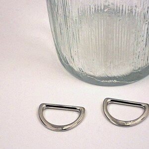 Puede incluir: Dos anillos en D de metal plateado, tambi&eacute;n conocidos como anillos en D o anillos en forma de D, se muestran sobre una superficie blanca. Los anillos tienen un di&aacute;metro de aproximadamente 2,5 cm.