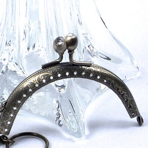 May include: A vintage-style, bronze-toned purse frame with a decorative floral pattern. The frame has a curved shape, with two small, clear, gem-like accents at the top. A small chain and ring are attached to one side. The frame is likely for a small bag or clutch.