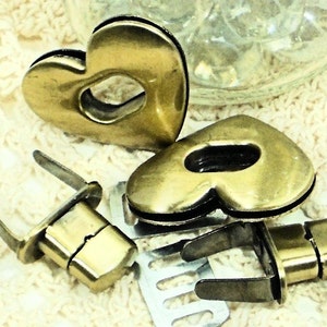 May include: Three gold-toned heart-shaped metal clasps with a hole in the center. Two of the clasps are larger and have a more rounded shape, while the third is smaller and more rectangular. The clasps are on a white crocheted surface.