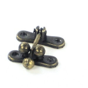 May include: Close-up of two antique bronze-colored clasps. Each clasp has a rectangular base with rounded ends and a small ball on each side. The clasps are designed to secure a box or other small container.