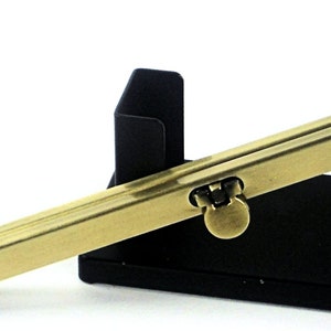 May include: A gold metal clasp with a black metal frame. The clasp has a decorative design and is used to secure a bag or purse.