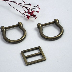 May include: Two antique gold-toned metal D-rings and a rectangular metal buckle. The D-rings are approximately 5 cm in diameter and the buckle is about 2.5 cm wide.