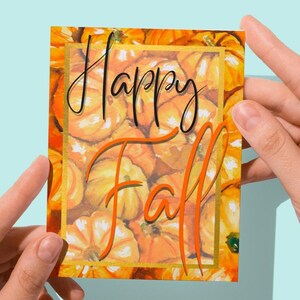 May include: A rectangular card with a watercolor design of pumpkins, with the words "Happy Fall" in black and orange script. The card is held by two hands against a light blue background.