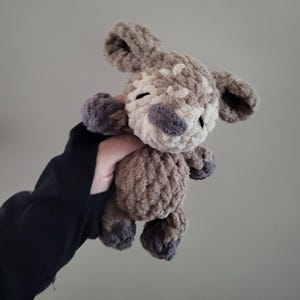 May include: A small, soft, crocheted plush animal toy made from textured, chunky yarn in shades of light brown, cream, and dark grey. The creature features large, rounded ears, a rounded snout, and dark grey accents on the paws and nose.
