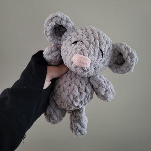 May include: A small, handmade crochet plush toy shaped like a mouse. The toy is crafted from soft, chunky grey yarn with a distinct textured stitch pattern. It features large, rounded ears, a small light pink nose, and simple black embroidered eyes.