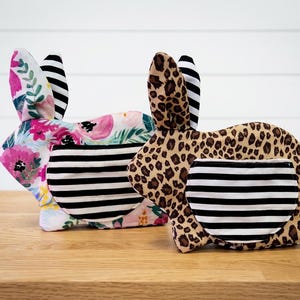 May include: Two stuffed rabbit-shaped fabric items. One features a pink and green floral pattern, the other a brown and black leopard print. Both have black and white horizontal striped pockets on the side and black and white striped inner ears.