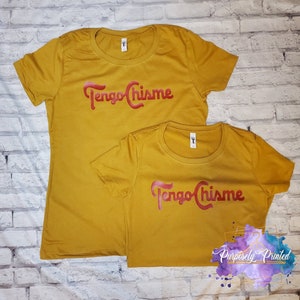 Puede incluir: Dos camisetas de color amarillo mostaza con la frase "Tengo Chismes" impresa en rojo en la parte delantera. Las camisetas son de diferentes tama&ntilde;os.