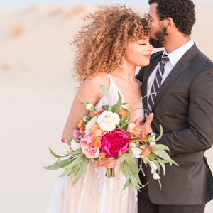 A colorful boho styled bridal bouquet is being held by a couple eloping in a sandy desert area. The artificial flowers include hot pink peony, pink and peach roses, white ranunculus and long leaf eucalyptus