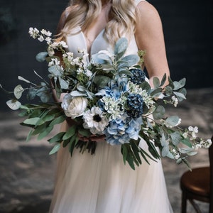 A blue and white flower bridal bouquet is being held by a lovely bride in a white dress in a dark blue room. The artificial flowers are complemented with various types of greenery including eucalyptus and lambs ear foliage