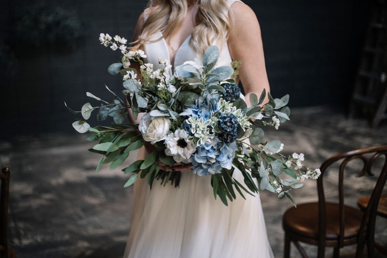 A blue and white flower bridal bouquet is being held by a lovely bride in a white dress in a dark blue room. The artificial flowers are complemented with various types of greenery including eucalyptus and lambs ear foliage