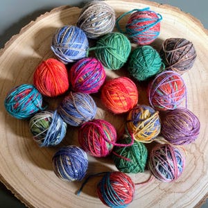 May include: A selection of multi-coloured yarn balls, including shades of blue, green, red, orange, pink, and purple. The yarn balls are displayed on a wooden surface, highlighting the diverse colours and textures. Perfect for knitting and crochet projects.