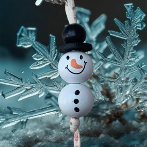 May include: A white and black snowman keyring with a black hat and a red nose. The snowman is made of wooden beads and has a white string attached to it. The keyring is hanging from a silver metal ring.