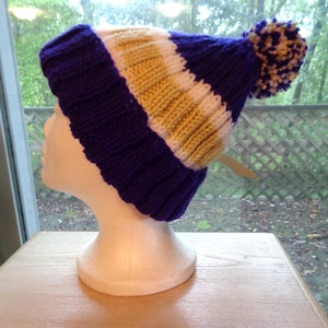 May include: A knit beanie featuring a ribbed purple cuff, followed by horizontal stripes of white and yellow yarn. The hat is topped with a multi-colored pom-pom made of purple, white, and yellow yarn, showcasing a thick, textured knit construction.