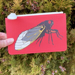 May include: A red zippered pouch featuring a detailed illustration of a cicada insect. The cicada has gray, yellow, and black markings. The pouch has a white zipper and a white pull tab. The background is a blurred view of green foliage.