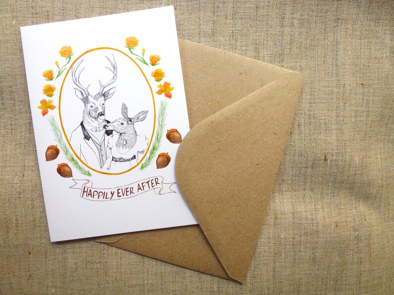 May include: A white card with a drawing of a deer couple in a gold oval frame. The deer are wearing clothes and are kissing. The card has orange flowers and brown acorns around the frame. The text "Happily Ever After" is written on a banner below the frame.