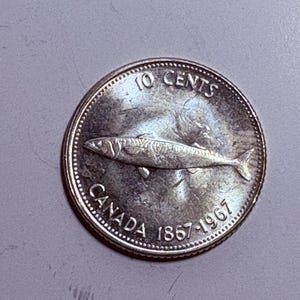 May include: A silver Canadian 10-cent coin from 1967. The coin features a fish in the centre, with the words "10 CENTS" at the top and "CANADA 1867-1967" around the bottom edge. The coin is round.