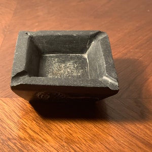 May include: A square, dark gray ashtray with a textured surface. The ashtray has a recessed interior and a small notch on one side. The ashtray is made of a solid material and sits on a wooden surface.