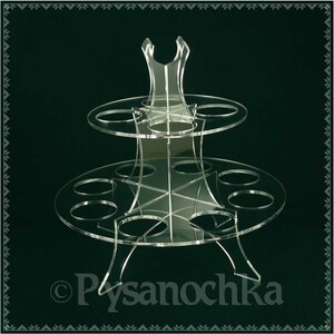 K&ouml;nnte beinhalten: Ein zweist&ouml;ckiger, transparenter Cupcake-St&auml;nder aus Acryl mit sechs runden Steckpl&auml;tzen auf jeder Ebene. Der St&auml;nder hat eine dekorative Oberseite mit einem Ausschnittmuster. Der St&auml;nder steht auf einem dunkelgr&uuml;nen Hintergrund.