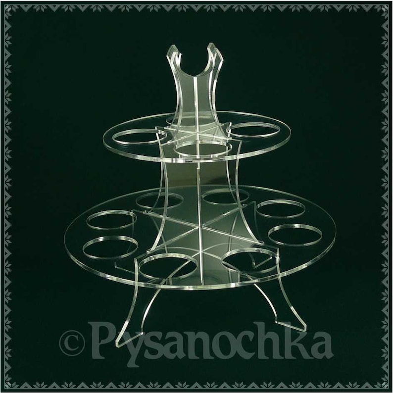 K&ouml;nnte beinhalten: Ein zweist&ouml;ckiger, transparenter Cupcake-St&auml;nder aus Acryl mit sechs runden Steckpl&auml;tzen auf jeder Ebene. Der St&auml;nder hat eine dekorative Oberseite mit einem Ausschnittmuster. Der St&auml;nder steht auf einem dunkelgr&uuml;nen Hintergrund.