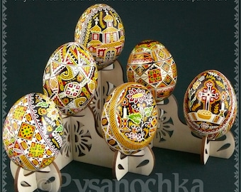 Puede incluir: Seis huevos de Pascua ucranianos pintados de forma intrincada, conocidos como pysanky, se exhiben en un soporte de madera. Los huevos presentan una variedad de diseños tradicionales, que incluyen patrones geométricos, motivos florales y símbolos religiosos. El soporte está hecho de madera de color claro y tiene un diseño recortado.