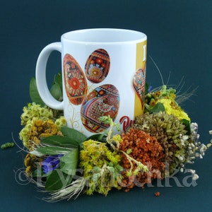 Peut inclure: Une tasse en céramique blanche avec un motif d'œufs de Pâques colorés et le texte "J'aime Pâques" sur le côté. La tasse est entourée d'une couronne de fleurs séchées.