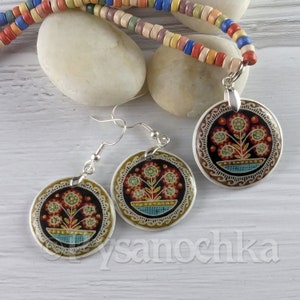 May include: A set of earrings and a necklace featuring a round pendant with a floral design. The pendant is painted with a colorful floral pattern on a dark background. The earrings and necklace are made of silver.