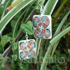 May include: A pair of square-shaped earrings with a white background and colorful geometric designs. The earrings feature a red, blue, and black pattern with white accents. The earrings are hanging from silver hooks.