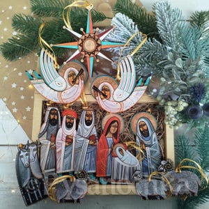 Op de afbeelding: Een houten kerstornamentenset met een ster, engelen en figuren uit de kerststal. De ornamenten zijn handbeschilderd met levendige kleuren en gouden accenten. De set wordt gepresenteerd in een houten kist en versierd met groen.