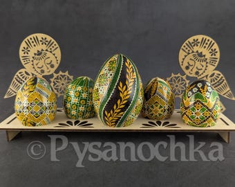Puede incluir: Expositor de madera con cinco huevos de Pascua decorados. Los huevos presentan patrones geométricos y florales en verde, amarillo y negro. Dos recortes de ángeles de madera flanquean los huevos, añadiendo un toque decorativo.