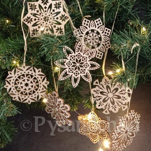 Puede incluir: Un conjunto de ocho adornos de madera en forma de copo de nieve con intrincados diseños cortados con láser. Los adornos están colgados de un árbol de Navidad con luces blancas.