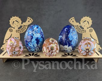 Puede incluir: Un expositor de madera con cinco huevos de Pascua decorados. Dos huevos azules tienen diseños florales, y uno tiene una escena nocturna. Dos huevos marrones tienen patrones florales. Dos recortes de ángeles de madera flanquean la exhibición. La palabra "Pysanochka" es visible.