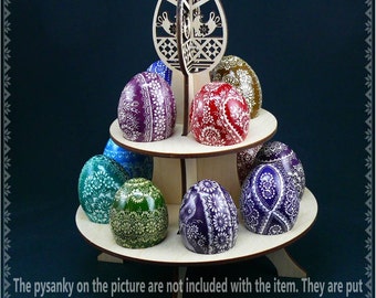 Puede incluir: Un soporte de madera de tres niveles para exhibir huevos de Pascua. El soporte tiene tres niveles y está decorado con un diseño recortado de un pájaro y un patrón floral. El soporte está vacío, pero hay huevos de Pascua decorados en el fondo.