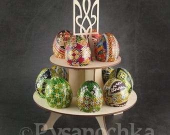 Puede incluir: Un soporte de madera con un diseño de tridente ucraniano en la parte superior, que sostiene una colección de huevos de Pascua pintados de forma intrincada. Los huevos están decorados con varios patrones y colores, incluyendo rojo, verde, amarillo y azul.