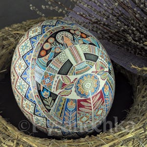 Può includere: Un uovo di Pasqua dipinto a mano con intricati motivi geometrici e una figura stilizzata al centro. L'uovo è decorato in tonalità di blu, rosso, giallo e verde, ed è annidato in un letto di paglia.