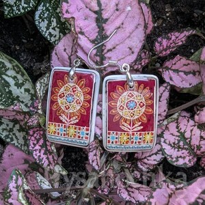 May include: A pair of rectangular dangle earrings featuring a deep red background with a central stylized sunflower motif in orange, yellow, and blue. The design includes a geometric border pattern at the base and silver-toned metal ear wires.