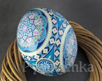 May include: A decorative, hand-painted egg with intricate geometric patterns in shades of blue, pink, and white. The egg is adorned with floral and diamond-shaped designs, resting on a woven, brown base. The egg's surface is glossy.