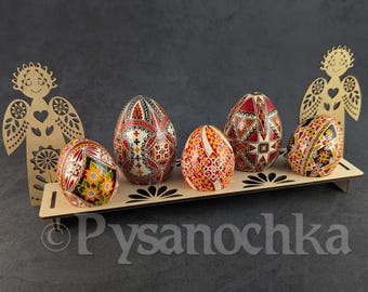 Puede incluir: Seis huevos de Pascua decorados con intrincados patrones en rojo, negro, amarillo y blanco descansan sobre una bandeja de madera. Dos recortes de ángeles de madera flanquean la bandeja. La palabra "Pysanochka" es visible.