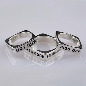 Puede incluir: Tres anillos de plata con caras cuadradas, cada uno grabado con una frase diferente en letras negras: "NOT NOW", "LIFE'S GOOD SEEKING LOVE" y "PISS OFF". Los anillos se muestran sobre una superficie reflectante.