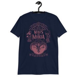 May include: Navy blue t-shirt featuring a pink graphic design with the text "MINES OF MORIA" and illustrations of Orcs, Goblins, and a Cave Troll. The design includes the text "Visit the lost kingdom of Khazad-dûm" and "Middle Earth Famous".
