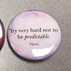 May include: A round button with a watercolor background in shades of purple and pink. The button features a quote in black text: "Try very hard not to be predictable." The quote is attributed to "Newt".