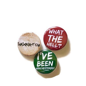 May include: Three round button pins with white text on colored backgrounds. The first pin is beige with the text "Sasquatch". The second pin is red with the text "What the hell?" The third pin is green with the text "I've been Winchestered!"
