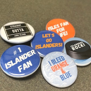May include: Six different button pins with various designs and text related to the New York Islanders hockey team. The pins are blue, orange, white, and black.