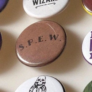 May include: A brown button with the text "S.P.E.W." in black lettering. The button is a round, flat, and has a glossy finish.