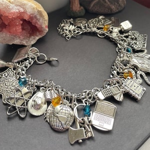 May include: Silver charm bracelet with various charms, including a globe, laptop, cowboy hat, and atomic symbol. The bracelet has a lobster claw clasp and several colorful beads. The charms are detailed and have an antique finish.