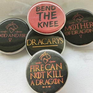 May include: Five round black and gold pins with different Game of Thrones quotes. The pins feature the phrases "Blood and Fire", "Bend the Knee", "Dracarys", "Mother of Dragons", and "Fire Can Not Kill a Dragon".
